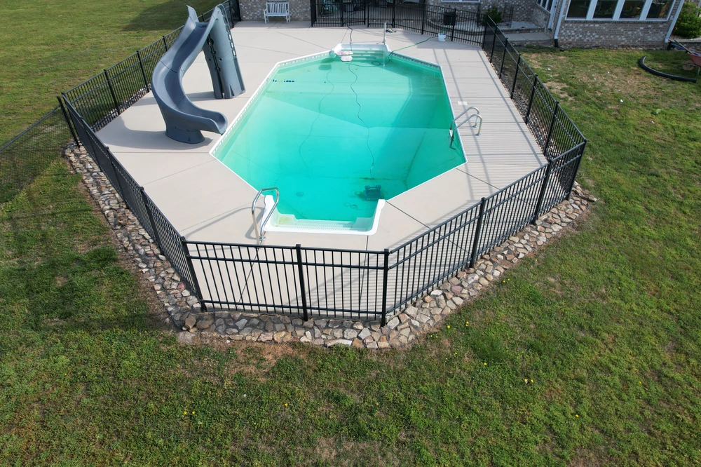 Swimming pool with concrete deck. Black fence. Rock border. Gray slides in a grassy backyard.