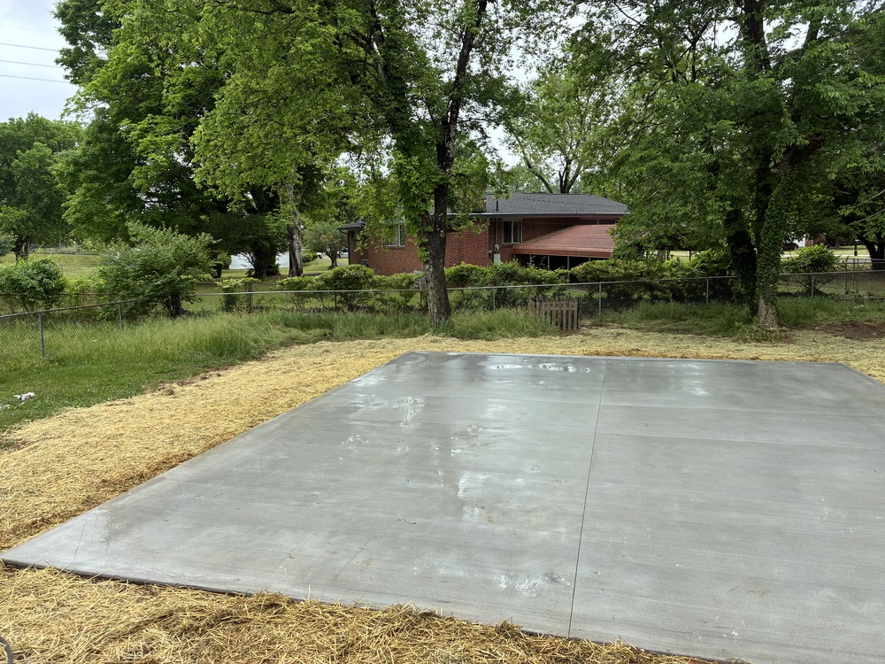 Large concrete slab bordered by straw in a backyard with trees and a fence.