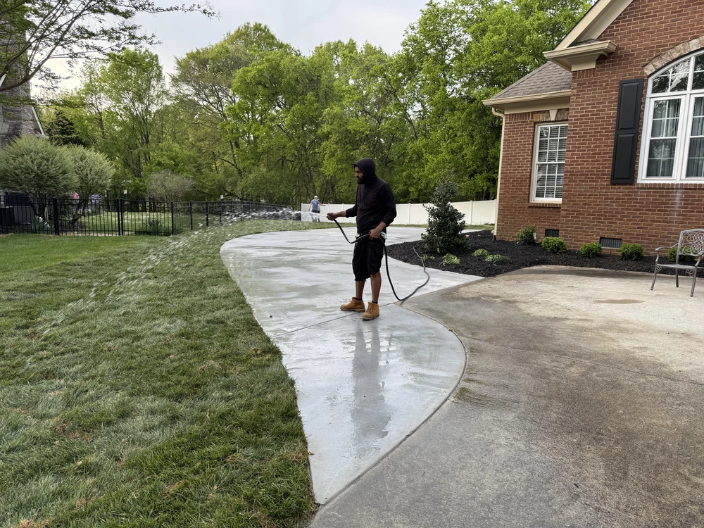 Worker rinses a new concrete driveway beside a brick house and landscaped yard.