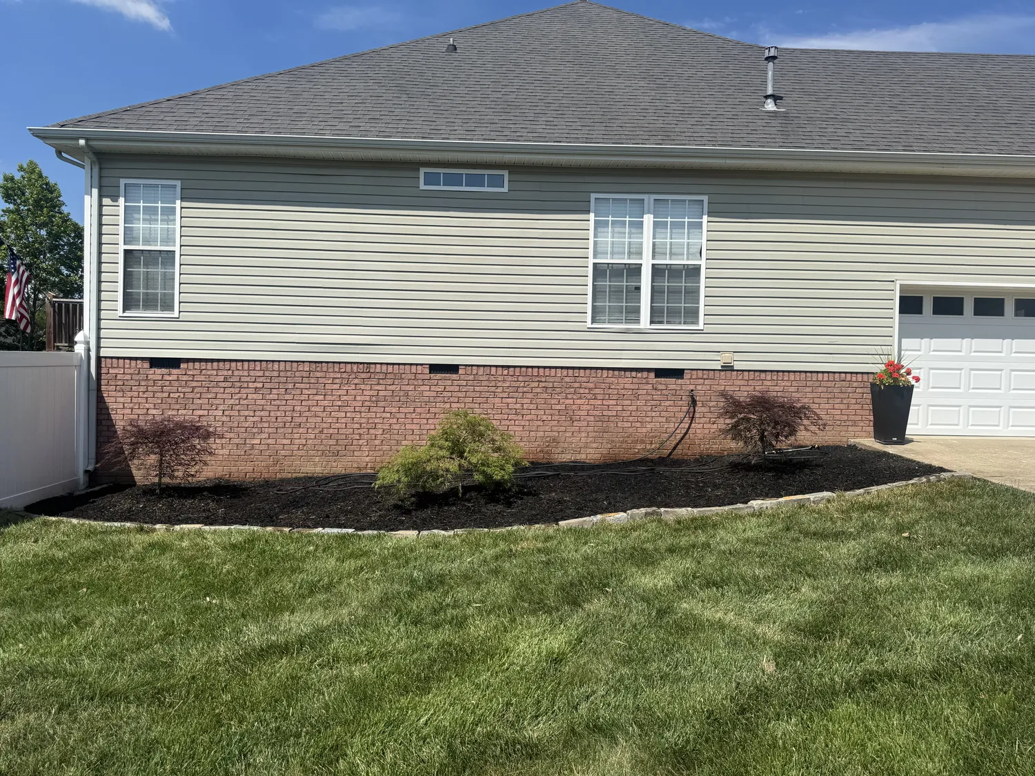 Young evergreens punctuate fresh black mulch along a sweeping retaining wall below the gray hillside home