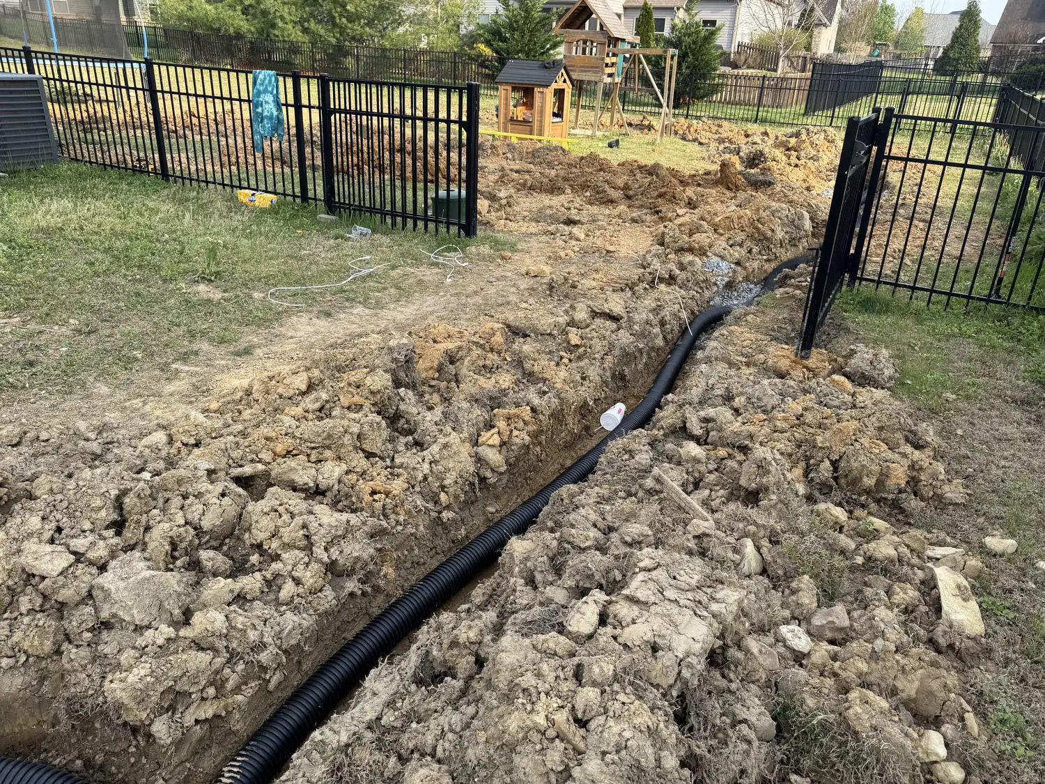 Fresh trenching carves through the fenced backyard as a black drain pipe snakes through rough soil toward better yard drainage