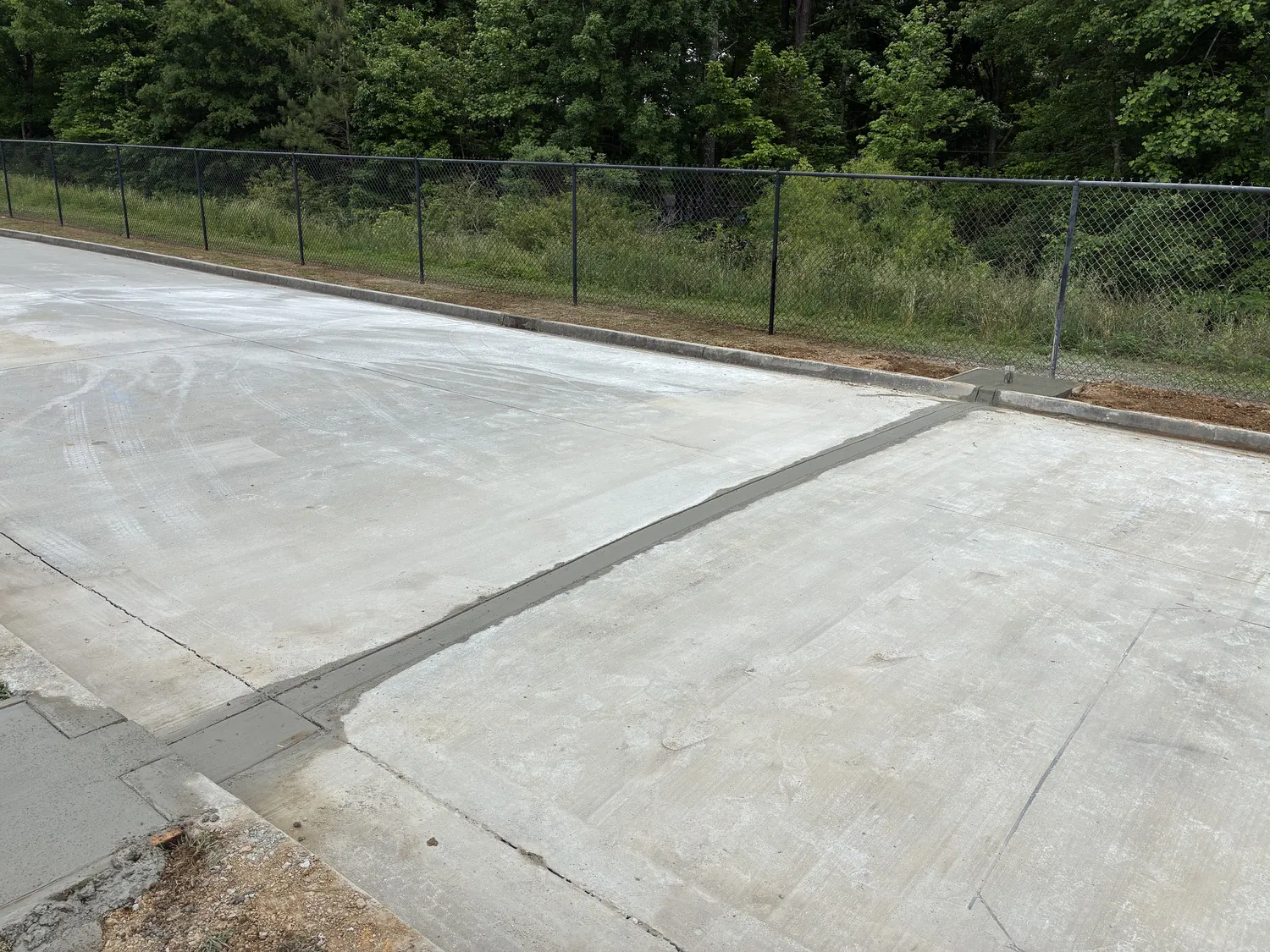 Bright concrete surface with narrow drainage channel, black chain-link fence, curb, and wooded area in the background