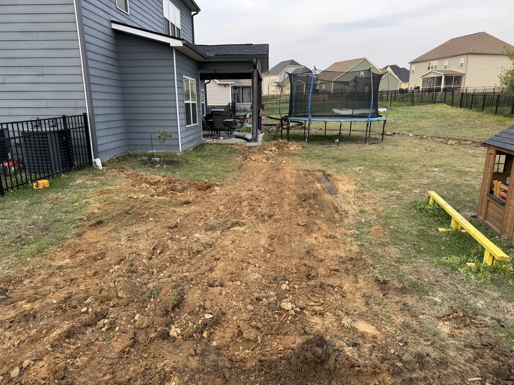 Fresh trench lines cut through the backyard as French drains take shape beside the gray home and trampoline