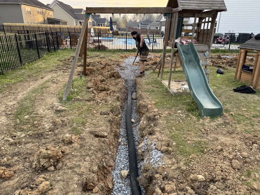 Backyard French drain installation with gravel black pipe and dug soil beside a slide in Cleveland, TN.