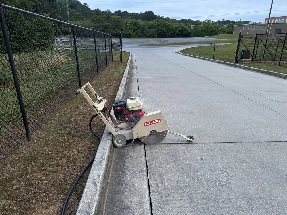 Walk behind concrete saw parked by a curb cut drainage line near a black fence in Cleveland, TN.