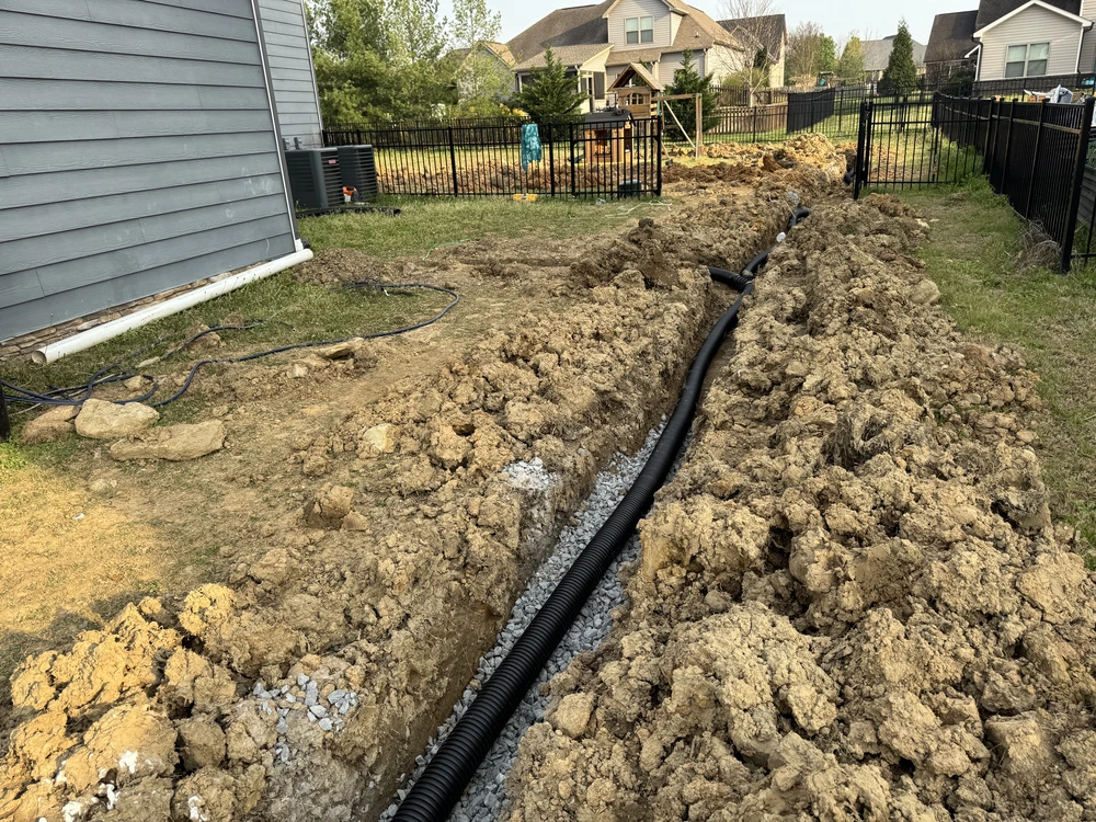 French drain trench with black corrugated pipe and gravel across a fenced backyard in Apison TN.