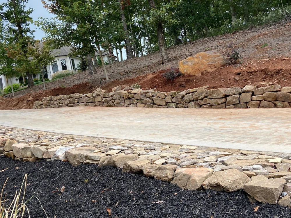 Smooth concrete walkway stretches below a rustic stone retaining wall, giving the wooded hillside a clean finished edge