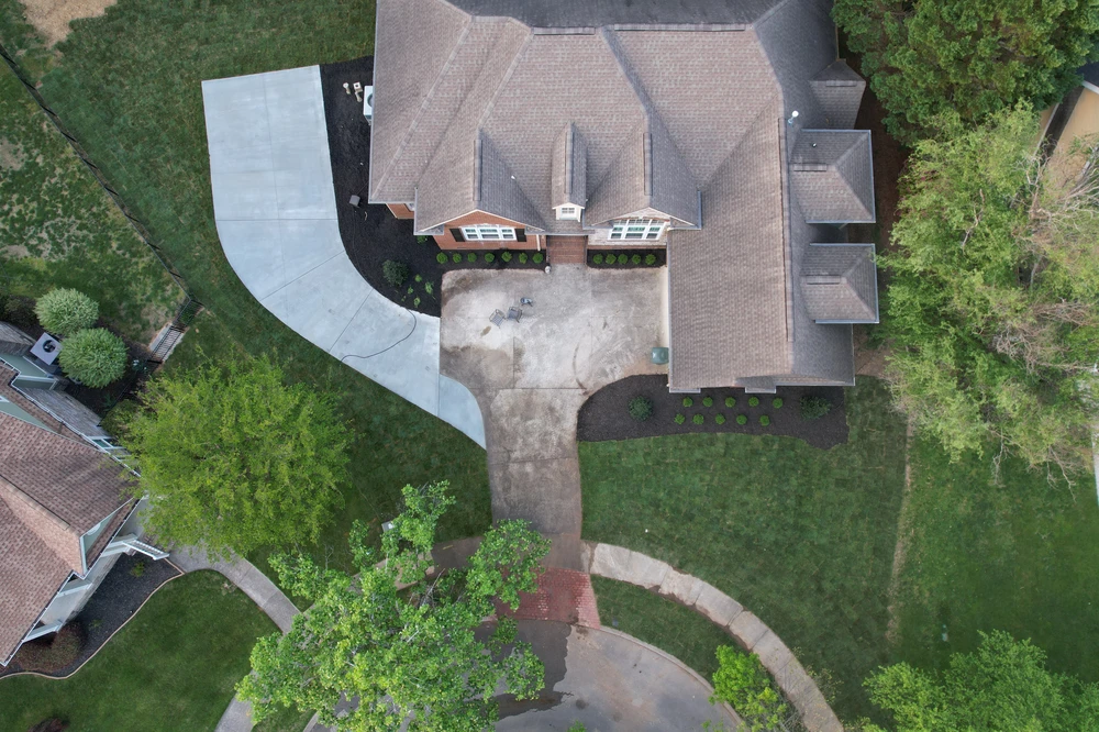 Aerial view of a brick home with curved concrete driveway black mulch beds and green lawn framing outdoor living spaces.