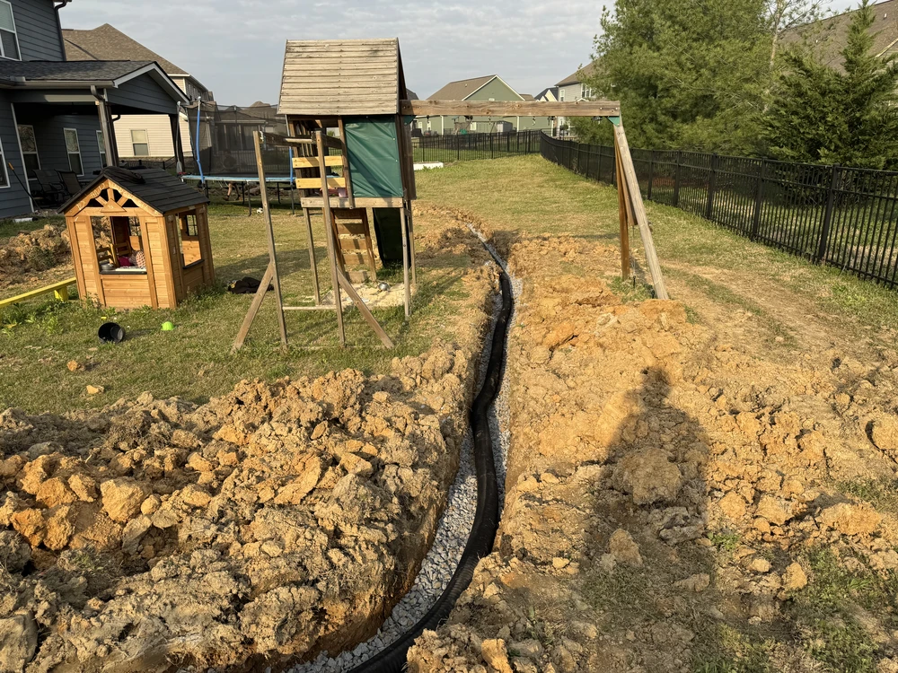 Chattanooga, TN backyard trench cuts past a playset as black drain pipe is set through sunlit clay for better runoff control