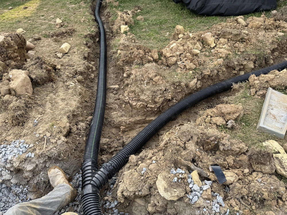 Black corrugated pipe curves through open clay trenches as French drains are laid beneath the yard