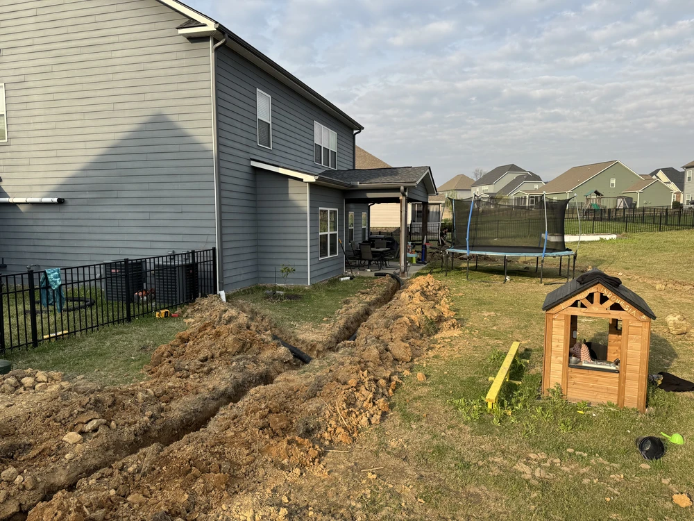 Ooltewah, TN backyard trench cuts through clay soil as drainage work reshapes the lawn beside a patio, playhouse, and trampoline