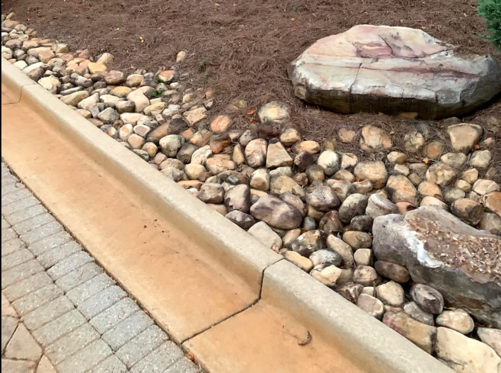 Rounded river rocks shape a clean yard drainage bed beside the curb, with pine straw and a flat boulder adding natural texture