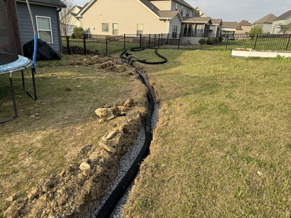 A long black pipe follows a curved backyard trench, guiding French drains across the sunlit lawn toward the fence