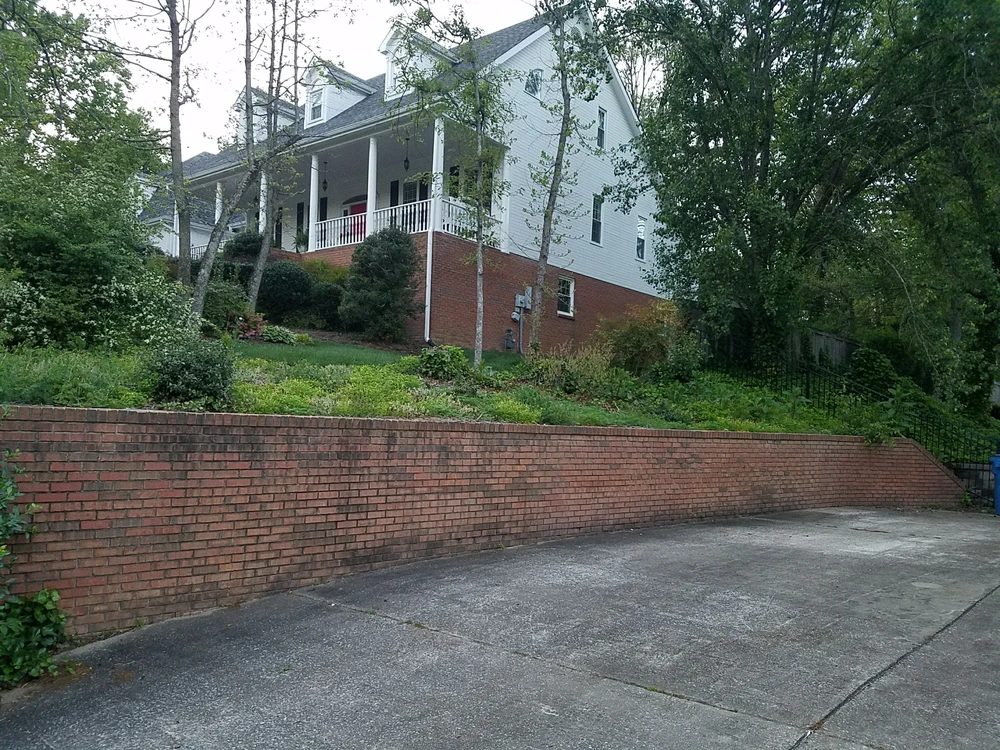 Young evergreens punctuate fresh black mulch along a sweeping retaining wall below the gray hillside home