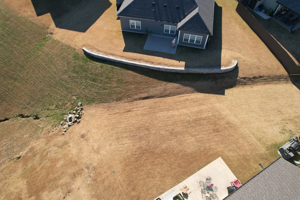 Curved concrete retaining wall behind a gray house holding back a dry sloped lawn from an aerial view.