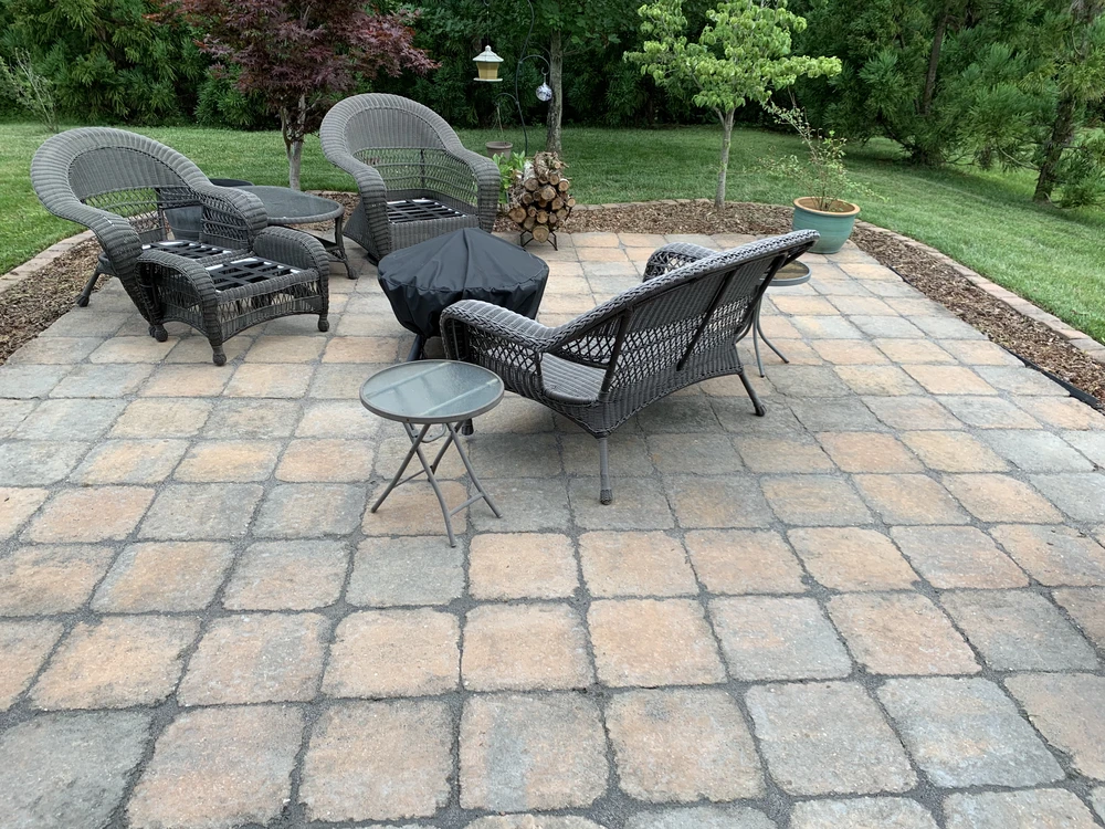 Paver patio with wicker seating fire pit and side tables overlooking a landscaped green yard.