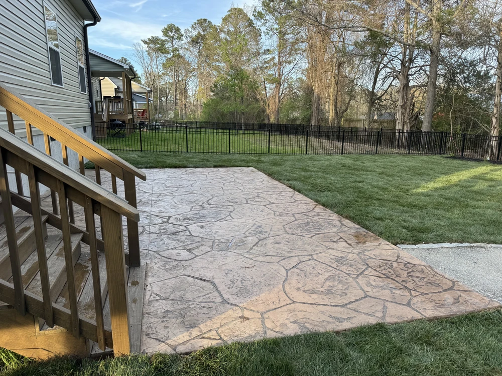 Stamped concrete patio beside wood deck steps with a fenced green backyard and fresh grass.