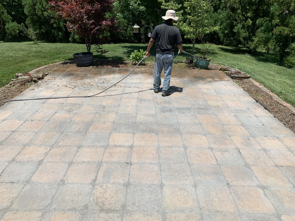 Worker pressure washes tan and gray pavers across a large backyard patio surrounded by grass.