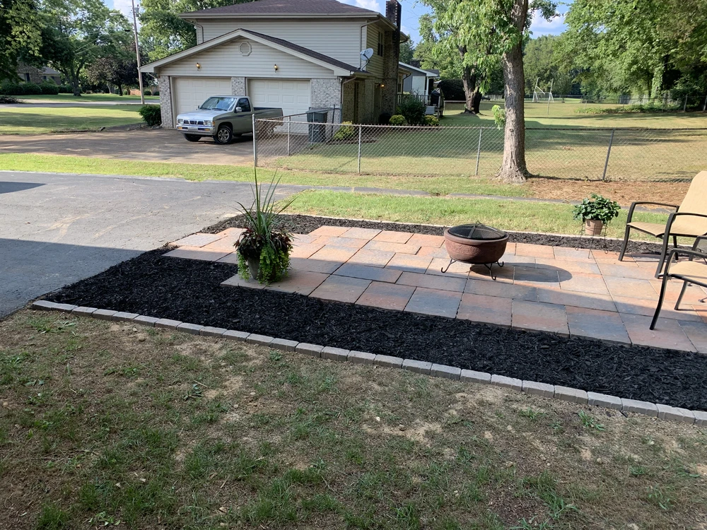 Tan pavers create a backyard patio edged with black mulch beside lawn chairs and a fire bowl in a fenced yard.