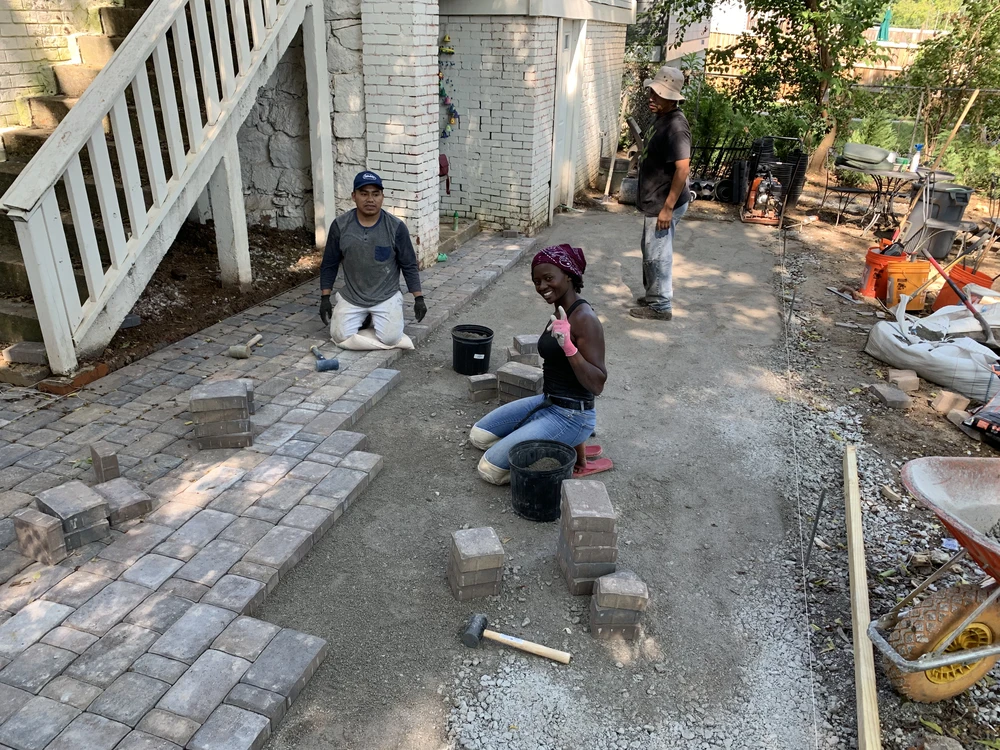 Three workers build brick patios beside a white house with gray pavers laid on the left and tools scattered across the sunlit yard.