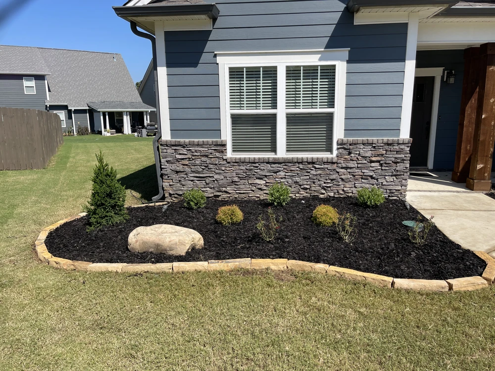 Fresh black mulch bed curves along a blue gray house with stone veneer shrubs and a tan border.