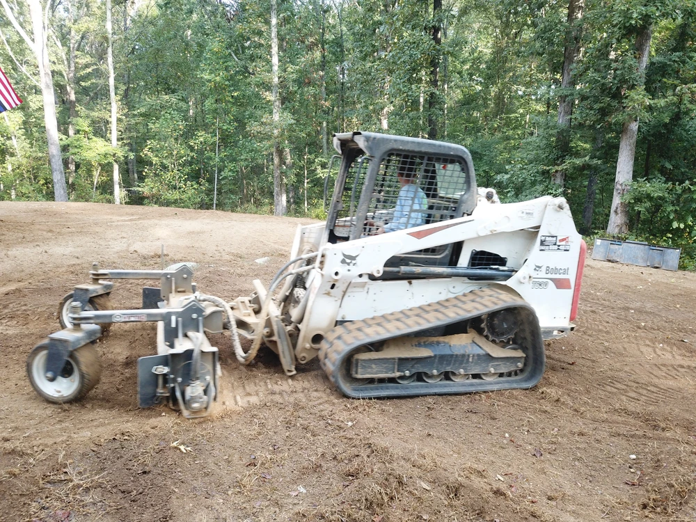 White Bobcat handles grading and resloping on a smooth tan pad at the edge of a dense green wooded lot.