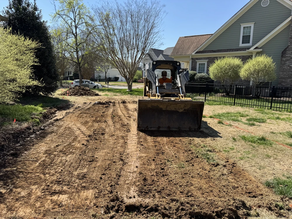 Skid steer grades a tan dirt path beside a gray green house with black fence bright shrubs and lawn.