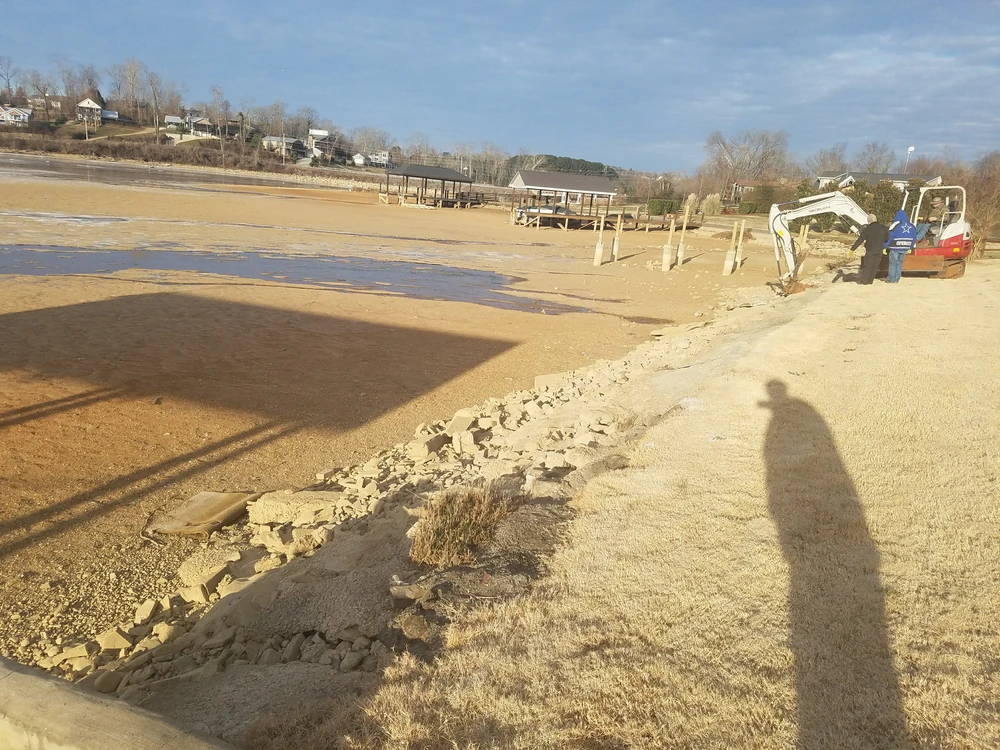 Excavation work shapes a sandy lakefront edge with rocks dry grass docks and pale water behind it.