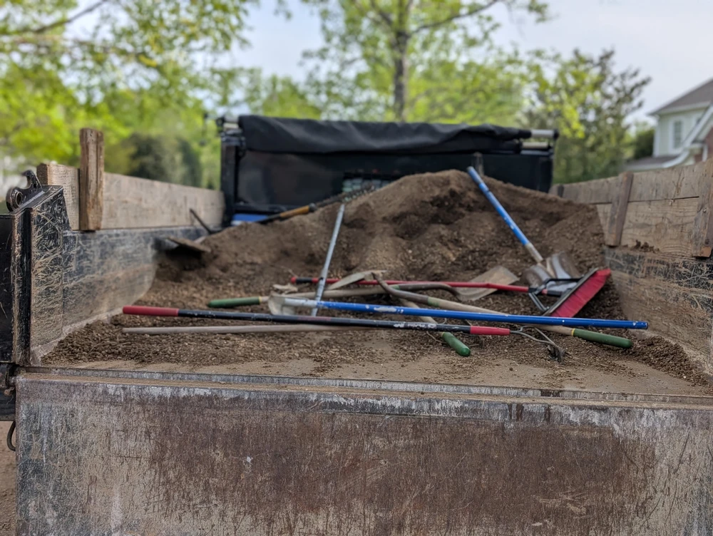 Shovels and rakes rest across a dirt pile in a wooden truck bed for an excavation project with green trees behind it.
