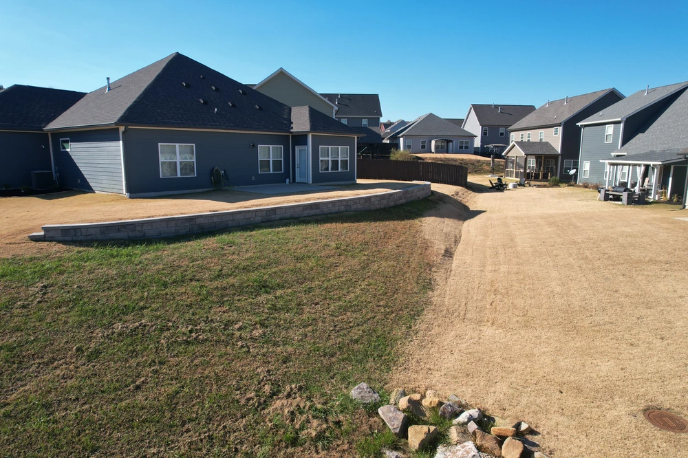 Curved gray hardscaping wall terraces a dry sloped backyard behind a blue gray house with neighboring homes beyond.
