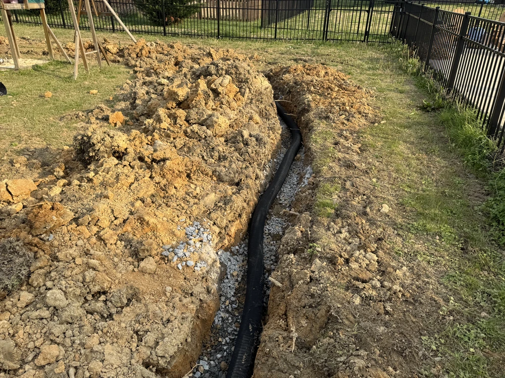French drain trench with black pipe and gravel through a fenced backyard under construction in Ringgold GA.