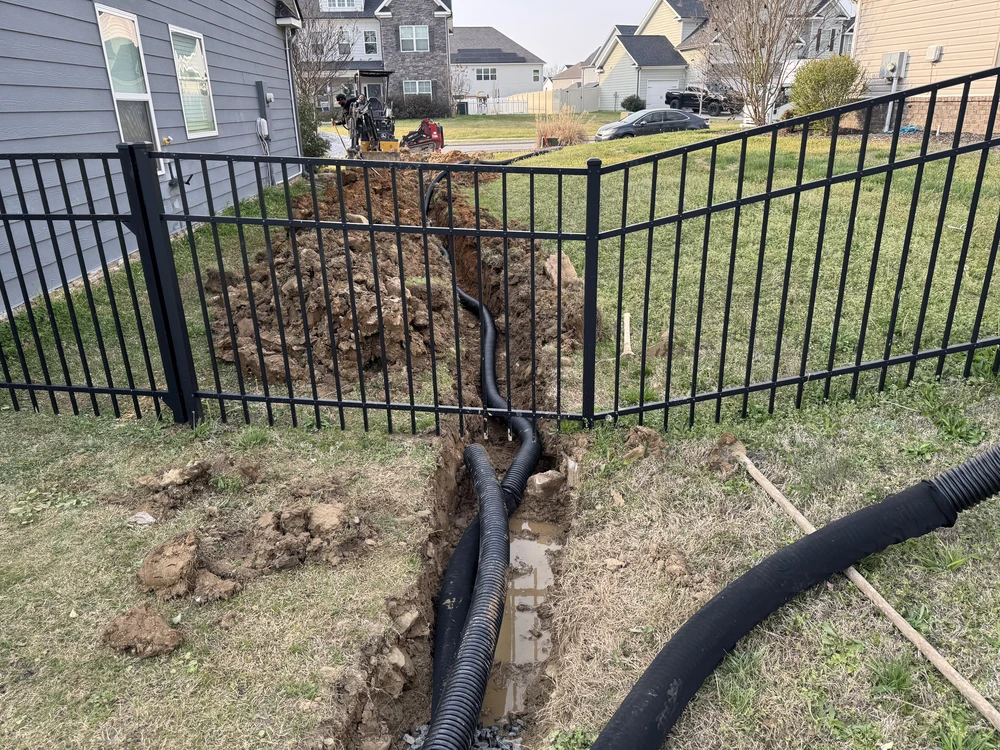 Black corrugated drain pipes run through a fenced yard trench beside a gray house in Red Bank TN.