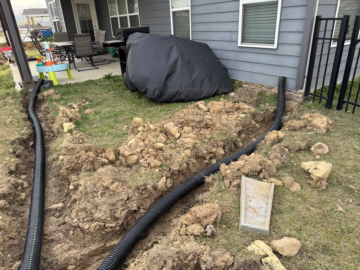 Trench drainage installation along a gray house patio with black corrugated pipe and exposed soil